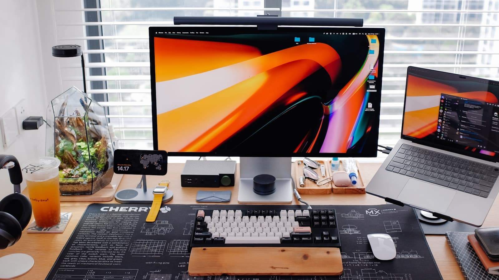 A desk with a large monitor, a laptop on a stand, and a mechanical keyboard